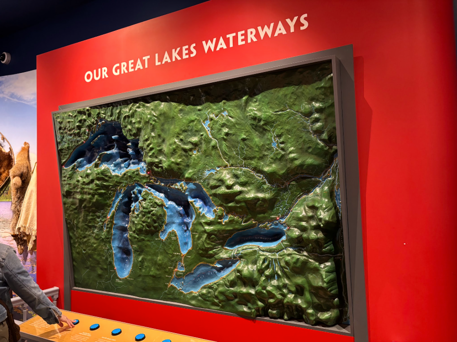a green and blue raised relief map of the Great Lakes hangs on a red wall. Below it, is a row of blue buttons, and a visitor is pressing one to light up several red points on the map