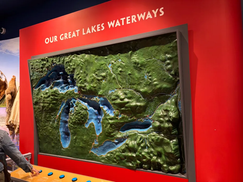 a green and blue raised relief map of the Great Lakes hangs on a red wall. Below it, is a row of blue buttons, and a visitor is pressing one to light up several red points on the map